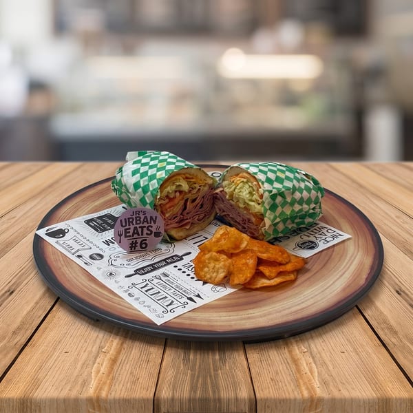 A JR's Urban Eats sandwich, cut in half, with chips on a wooden plate with branded paper.
