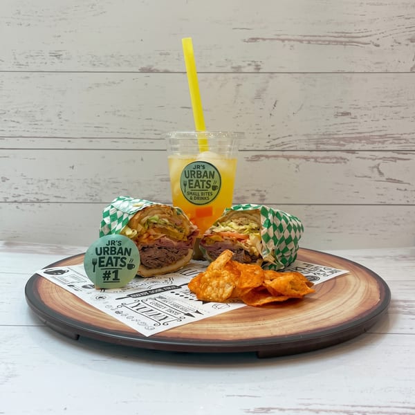 Two JR's Urban Eats sandwiches, chips, and a drink with a yellow straw on a wooden platter.